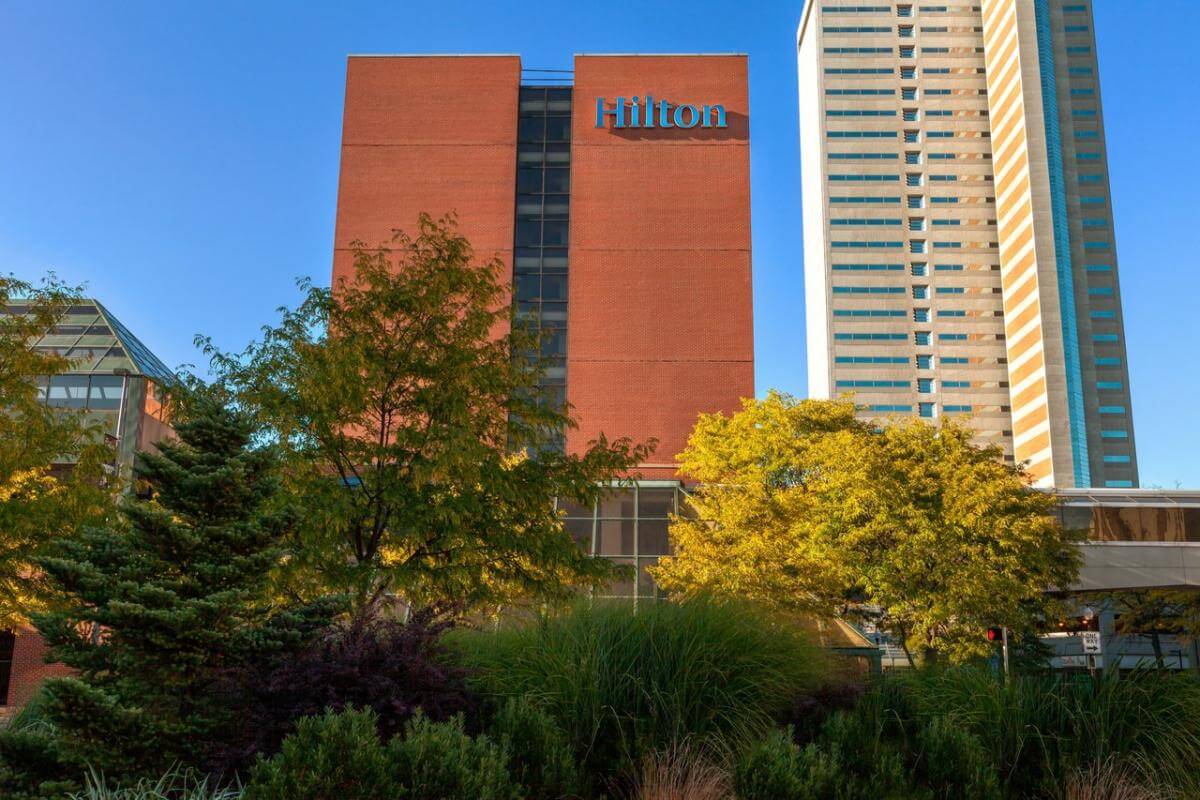 The image shows a Hilton hotel building with a red-brick facade and a tall, modern structure nearby. It is surrounded by lush green trees under a clear blue sky.